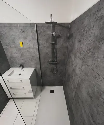 Même salle de bain après travaux : douche à paroi vitrée, colonne de douche, meuble vasque blanc et carrelage gris